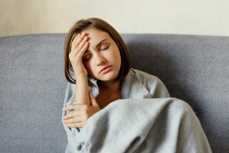 cold and stressed woman wrapped in blanket on couch with hand on forehead